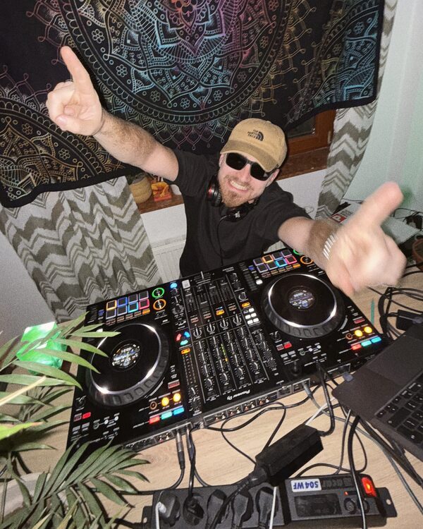 DJ w czapce i okularach przy konsolecie, za nim gobelin z mandalą